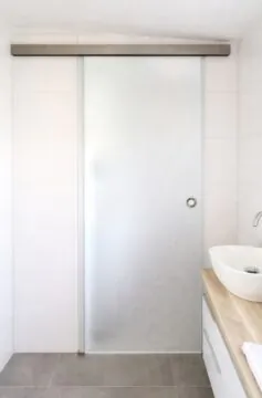 Fron­tale Ansicht einer satinierten Glasschiebetür im Badezimmer mit Edelstahl-Laufschiene und Griffmuschel.